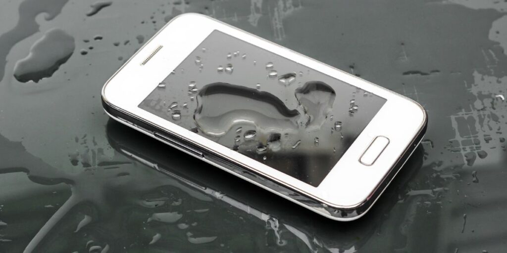 A smartphone with water droplets on its screen, sitting on a wet surface, indicating potential water exposure.