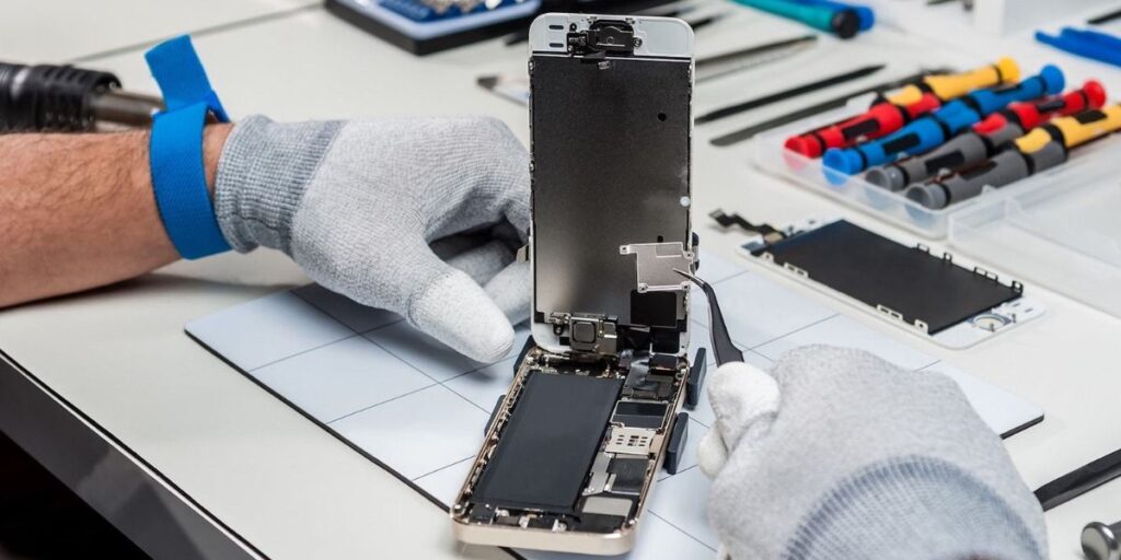 Gloved technician repairing an iPhone on a white workbench with tools and parts visible.