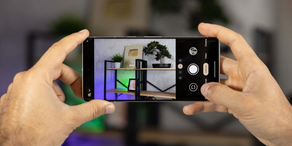 A pair of hands taking a picture with a smartphone camera of a shelf setup with a bonsai and YouTube award.