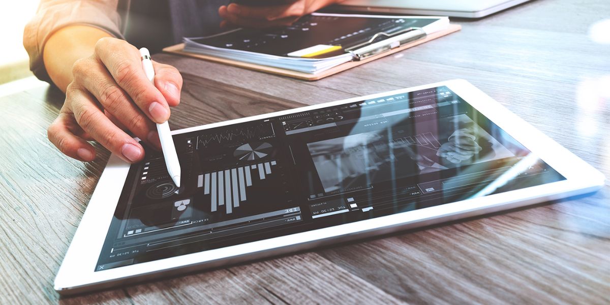 An executive uses a stylus on a white tablet displaying data analytics and charts on a wooden desk.
