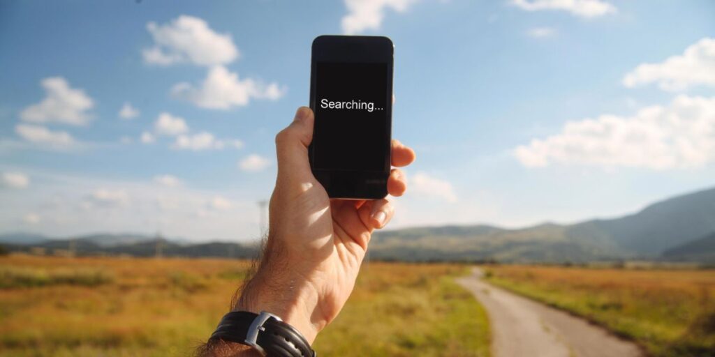 A hand holds a smartphone displaying Searching on a black screen in a rural, open landscape under a blue sky