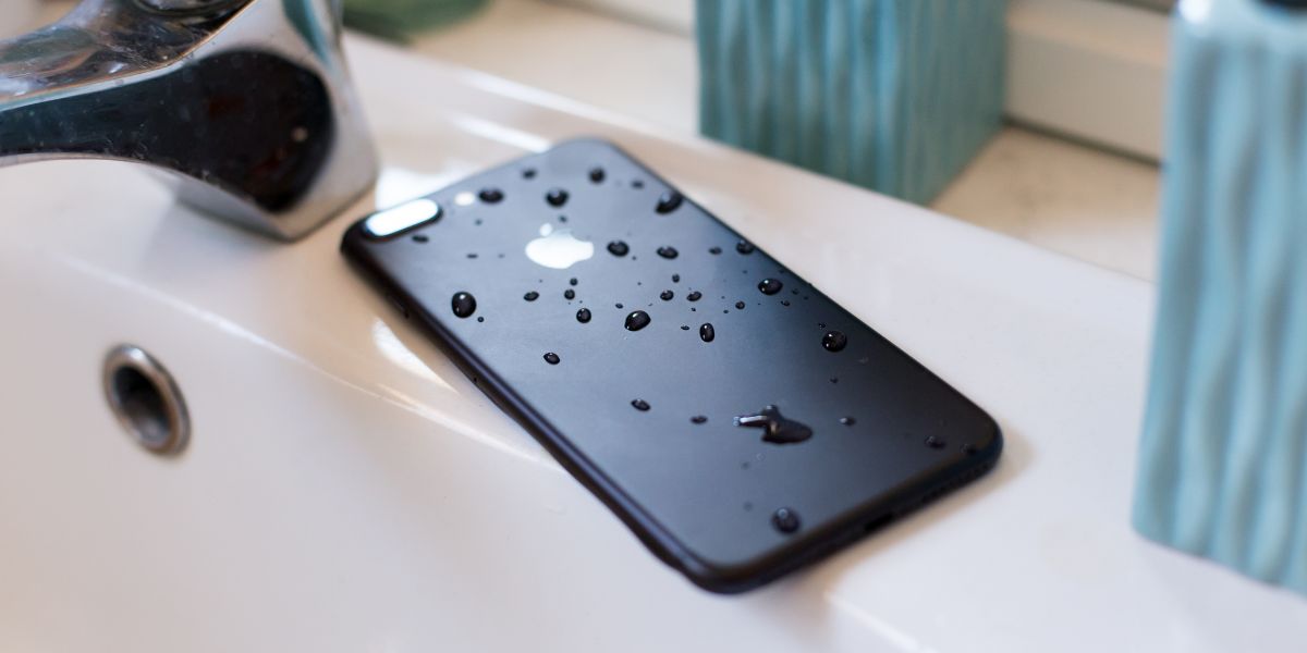 Black smartphone covered in water droplets lying in a white bathroom sink next to a faucet