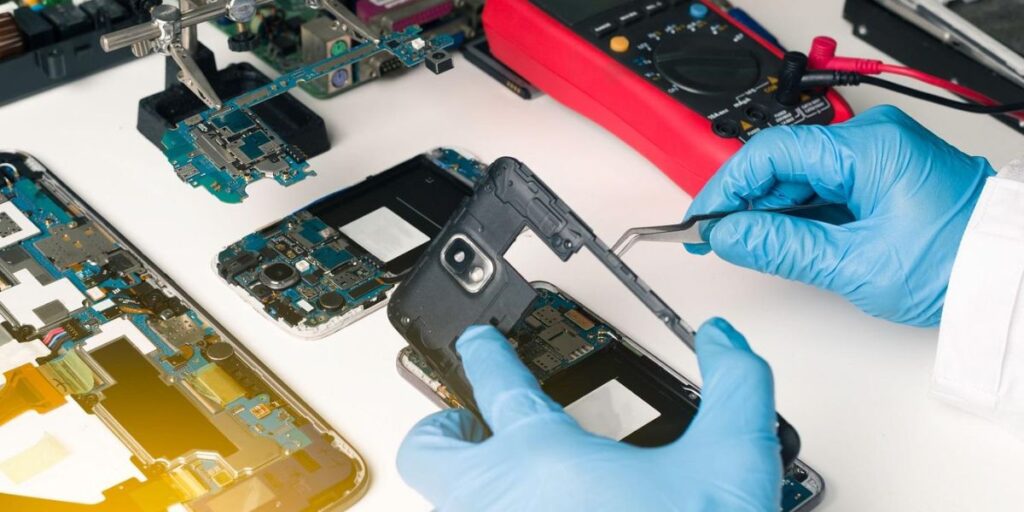 Gloved hands disassembling a smartphone for repair, with circuit boards and a multimeter visible on a desk