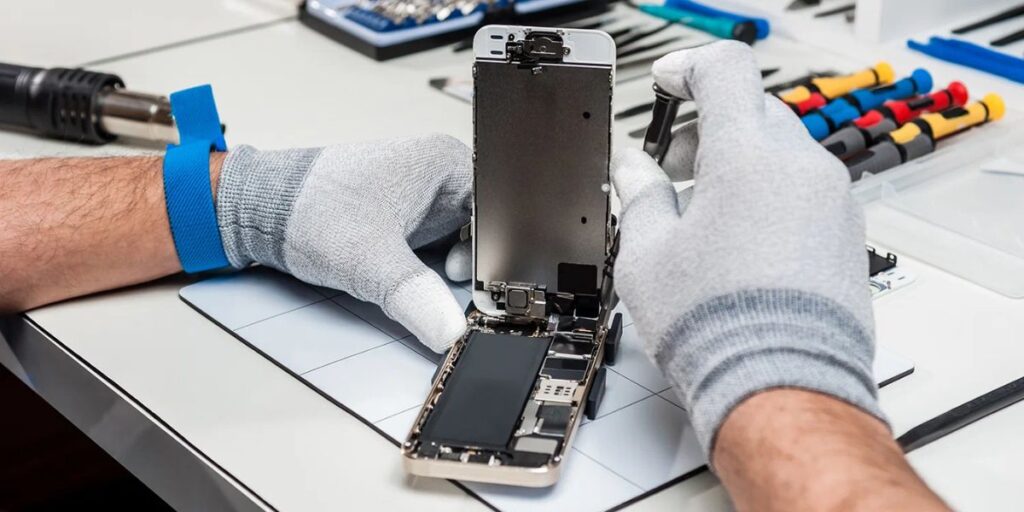 Hands wearing white gloves repairing an open smartphone on a workbench with tools nearby.