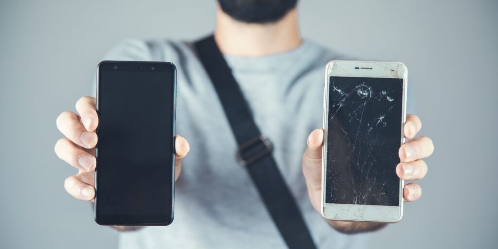 A person holds two smartphones; one has a perfectly clear black screen while the other has a badly cracked screen