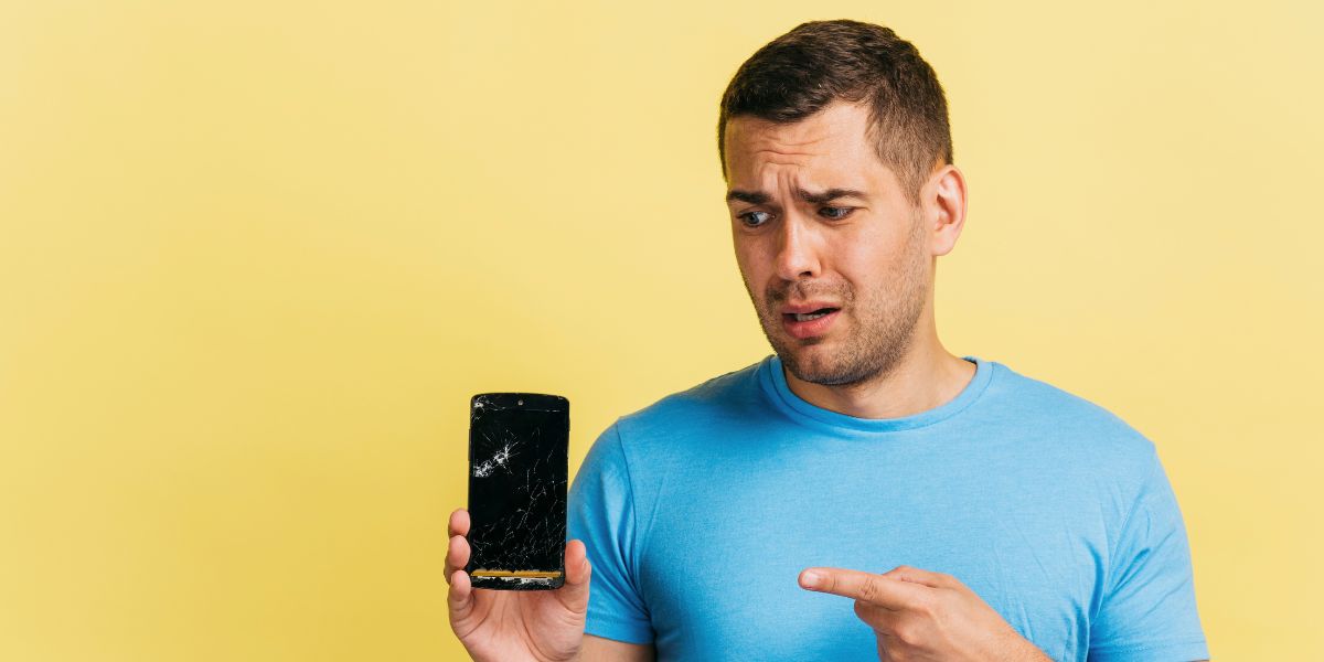A man in a blue shirt looks distressed while pointing at a smartphone with a shattered screen against a yellow wall